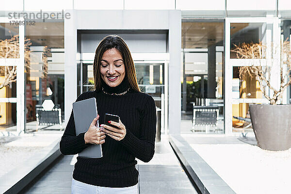 Glückliche Geschäftsfrau mit Laptop und Smartphone vor dem Café an einem sonnigen Tag