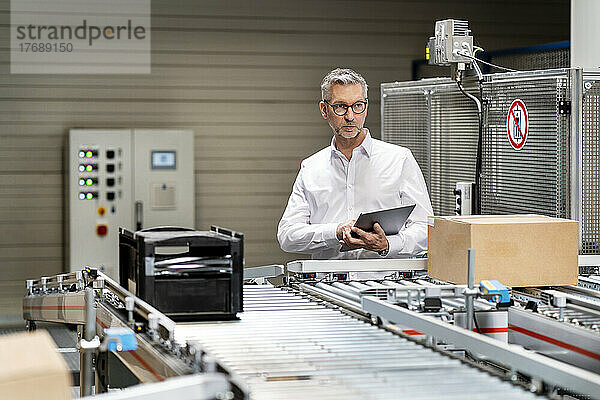 Reifer Geschäftsmann untersucht Karton auf Förderband mit Tablet-PC in der Fabrik
