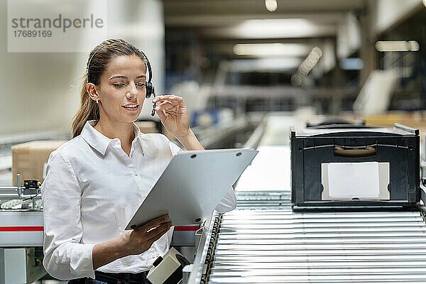 Junge Geschäftsfrau liest in der Fabrik mit Headset auf Klemmbrett