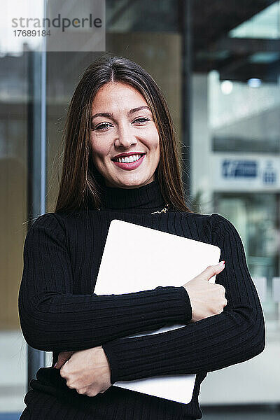 Lächelnde junge Geschäftsfrau mit Laptop steht vor einer Glaswand