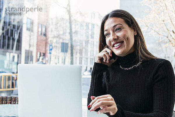 Lächelnde junge Geschäftsfrau mit Laptop  die im Café mit dem Handy telefoniert