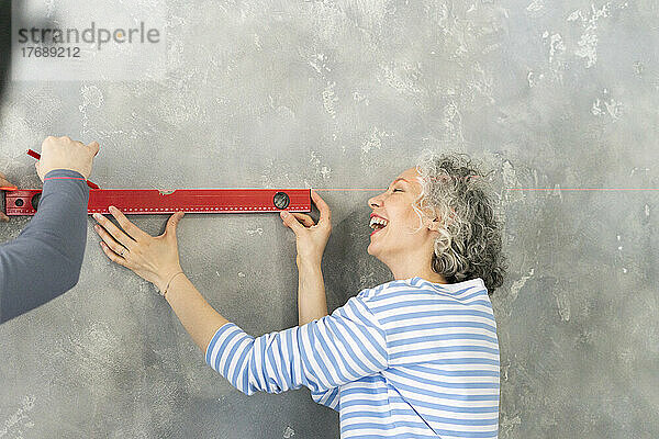 Glückliche Frau mit Freundin  die zu Hause ein Füllstandmessgerät an einer grauen Wand benutzt