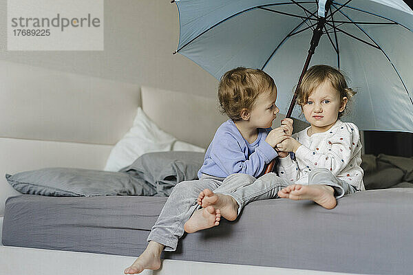 Twin sisters sitting with umbrella on bed at home
