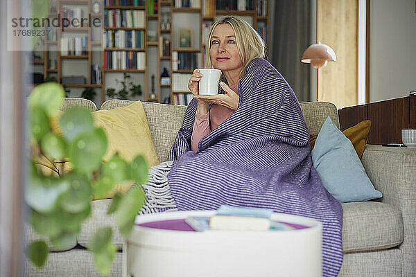 Blonde Frau  in eine Decke gehüllt  hält eine Kaffeetasse auf dem Sofa und träumt zu Hause
