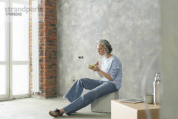 Frau isst Salat und sitzt auf einer Kiste vor der grauen Wand im neuen Zuhause