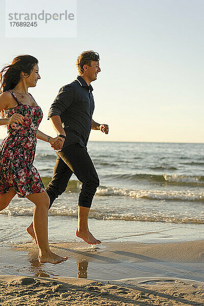 Glückliches Paar Händchen haltend beim Laufen am Strand an einem sonnigen Tag
