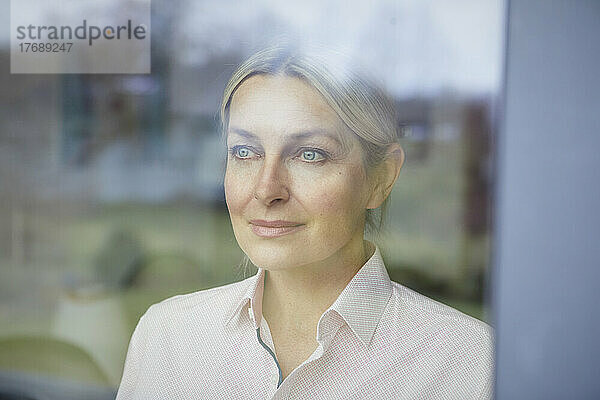 Lächelnde Frau mit blonden Haaren  die durch ein Glasfenster schaut