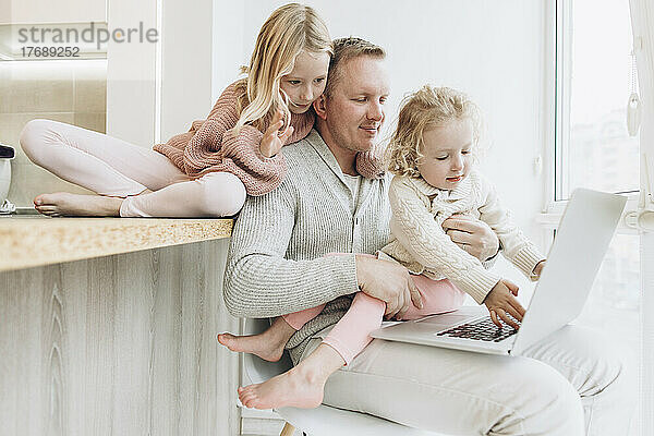 Töchter und Vater arbeiten am Laptop und sitzen zu Hause am Fenster