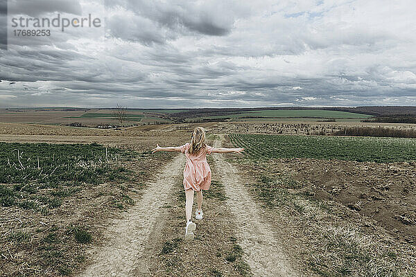 Sorgloses Mädchen läuft mit ausgestreckten Armen auf einem Weg in einem landwirtschaftlichen Feld