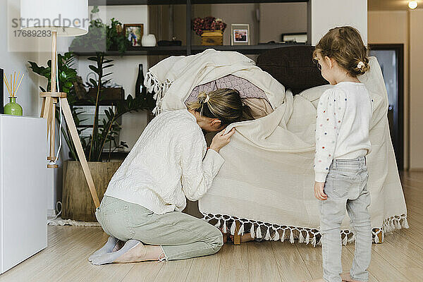 Playful mother peeking in blanket by daughter standing at home