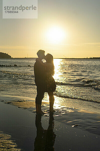 Young couple kissing each other in front of sea on beach at sunset