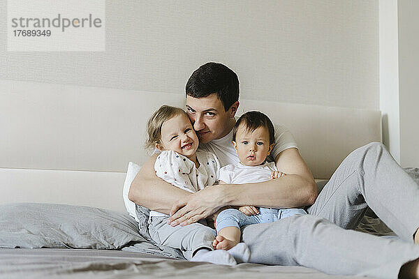 Lächelnder Mann umarmt Sohn und Tochter  die zu Hause im Bett sitzen