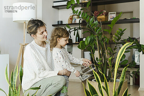 Girl watering potted plant with mother in living room