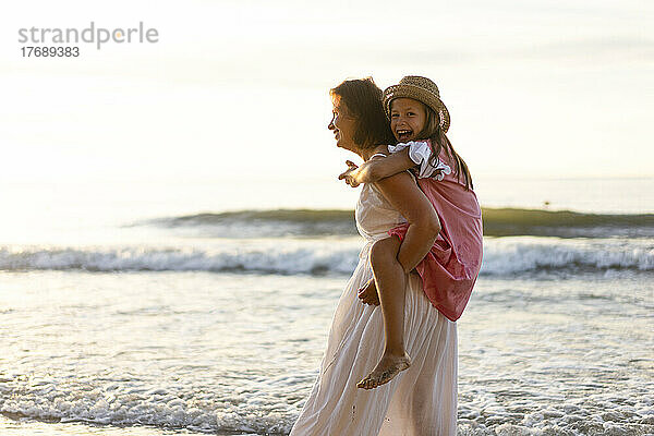 Lächelnde Mutter gibt fröhlicher Tochter Huckepackfahrt am Strand