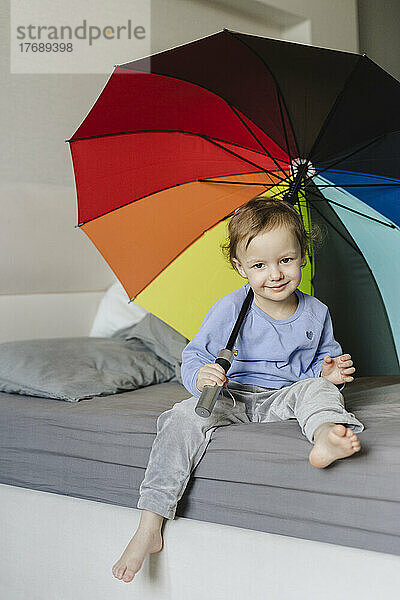 Lächelndes Mädchen  das zu Hause mit Regenschirm auf dem Bett sitzt