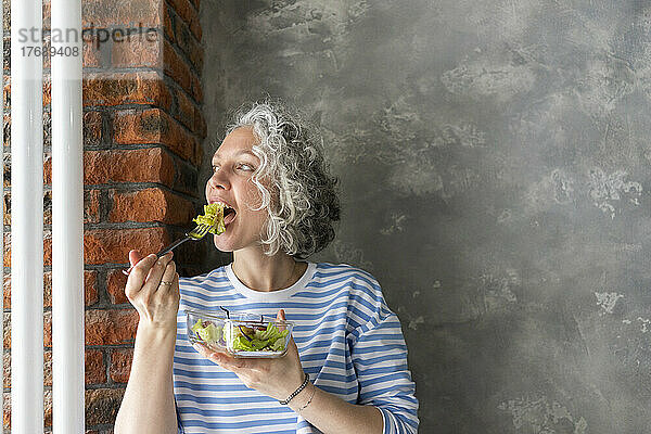 Frau isst frischen Salat zum Mittagessen vor grauer Wand