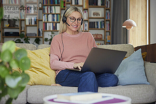 Glückliche Frau mit Headset und Laptop auf dem heimischen Sofa