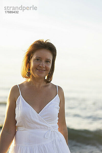 Lächelnde Frau steht im weißen Kleid am Strand