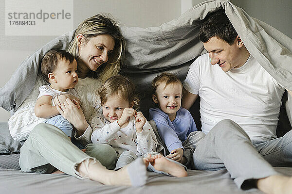 Glückliche Eltern mit Kindern  die unter einer Decke im Schlafzimmer sitzen