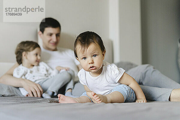 Niedlicher kleiner Junge mit Vater und Schwester im Hintergrund zu Hause im Bett