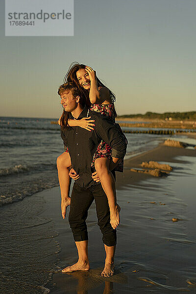 Junger Mann gibt Frau am Strand bei Sonnenuntergang Huckepackfahrt