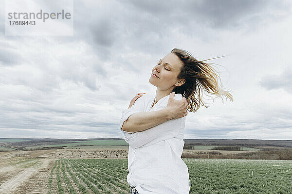 Frau mit geschlossenen Augen umarmt sich selbst auf einem landwirtschaftlichen Feld