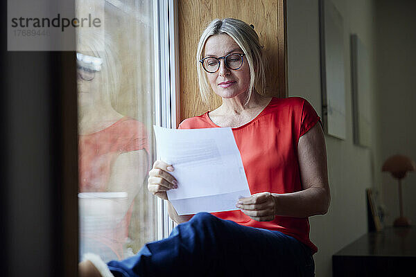 Reife Frau mit Brille liest Brief und sitzt zu Hause am Fenster