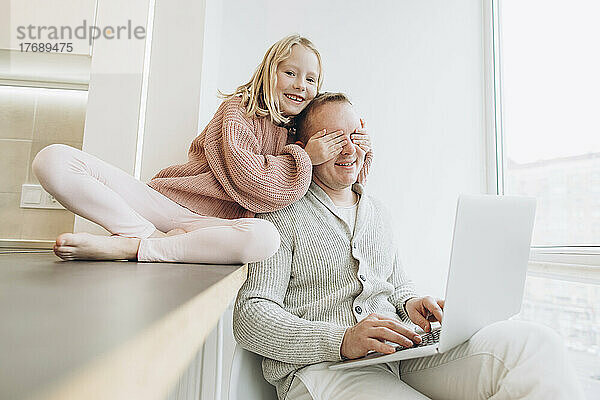 Glückliche Tochter bedeckt die Augen ihres Vaters  der zu Hause mit Laptop am Fenster sitzt