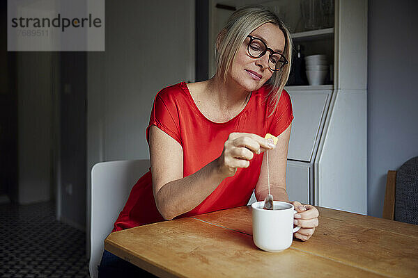 Reife Frau mit Teebeutel bereitet am Tisch Tee in einer Tasse zu