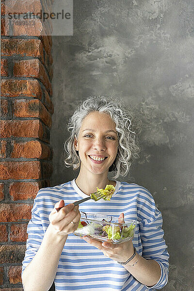 Glückliche Frau  die beim Mittagessen vor der Wand Salat isst