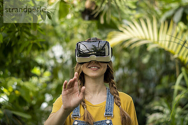 Lächelnde junge Frau mit Virtual-Reality-Simulator gestikuliert im Garten