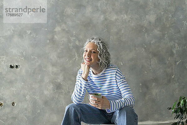 Glückliche Frau mit der Hand am Kinn und einer Tasse  die vor der Wand sitzt