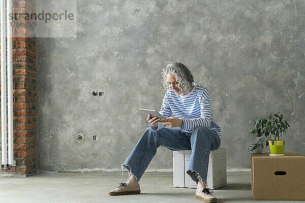 Lächelnde Frau mit grauen Haaren sitzt auf einem Kasten vor der Wand und benutzt einen Tablet-PC