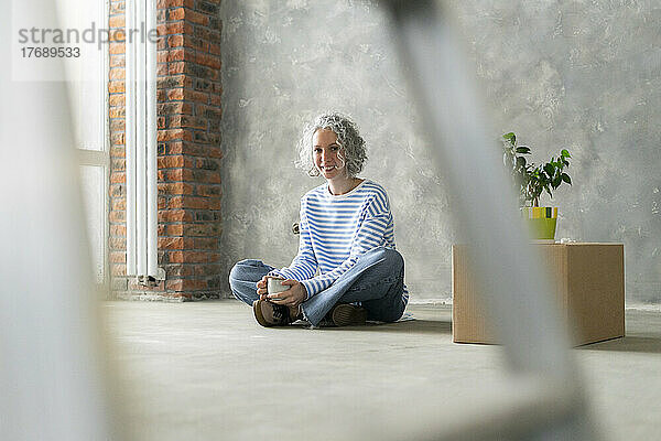 Glückliche Frau sitzt mit einer Tasse vor der Wand auf dem Boden