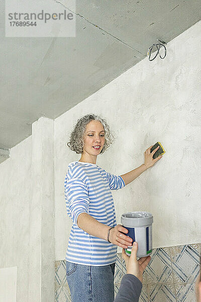 Lächelnde Frau mit Freundin schrubbt Wand im neuen Zuhause