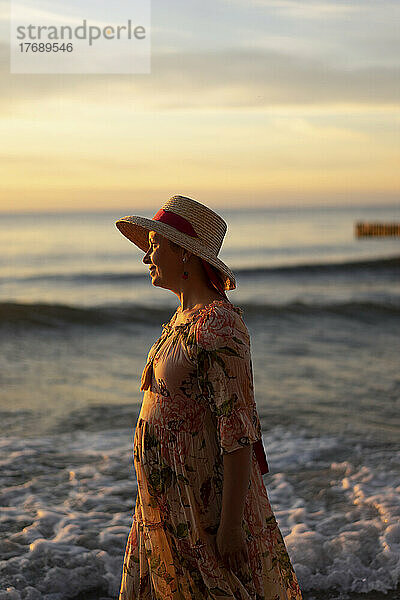 Frau mit Hut steht am Strand vor dem Meer