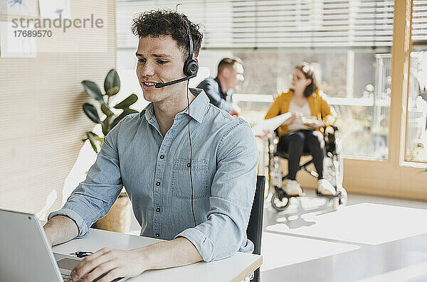 Junger Mann mit Headset und Laptop am Schreibtisch im Büro