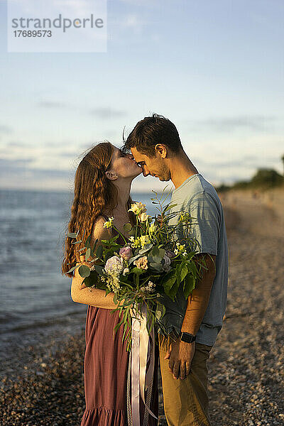 Woman holding bouquet kissing on man's forehead at beach