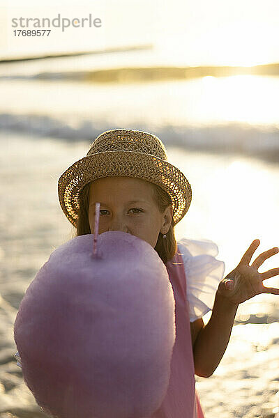Mädchen mit Hut isst an einem sonnigen Tag am Strand Zuckerwatte