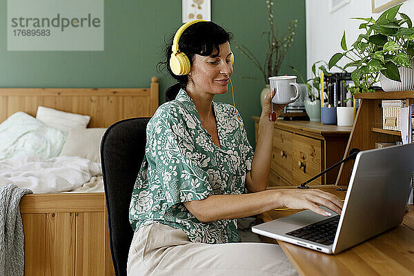 Lächelnde Geschäftsfrau  die zu Hause am Laptop Musik über Kopfhörer hört