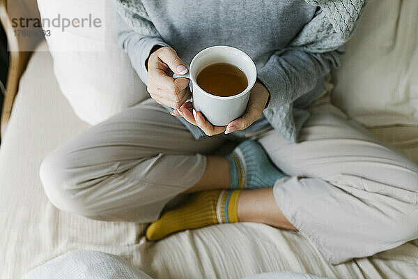 Hände einer Frau  die eine Teetasse hält und auf dem Bett sitzt