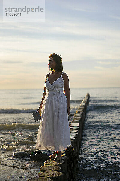 Lächelnde Frau  die bei Sonnenuntergang auf einem Holzpfosten vor dem Meer steht