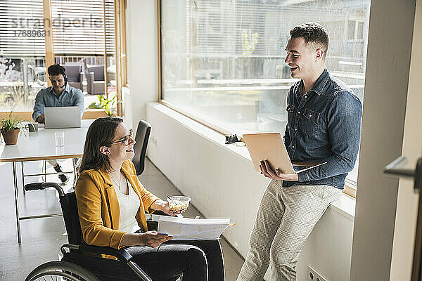 Lächelnder Geschäftsmann im Gespräch mit einer Kollegin im Rollstuhl im Büro