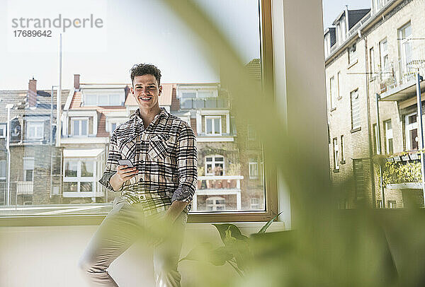 Porträt eines lächelnden jungen Geschäftsmannes mit Smartphone am Fenster im Büro