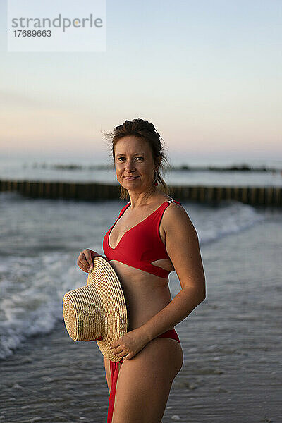 Frau im Bikini steht mit Hut am Strand