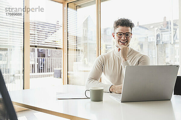 Lächelnder junger Geschäftsmann mit Laptop am Schreibtisch im Büro