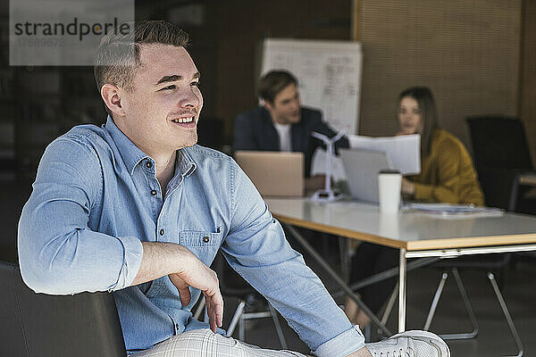 Selbstbewusster Geschäftsmann sitzt auf einem Stuhl im Büro mit Kollegen im Hintergrund