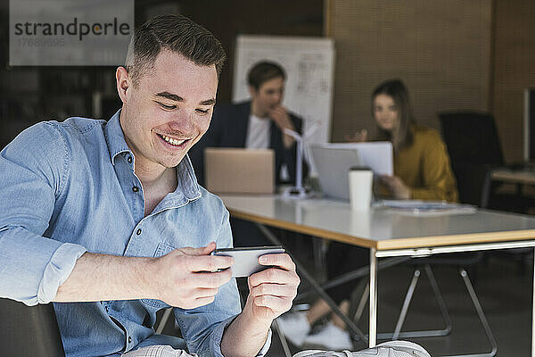 Lächelnder Geschäftsmann nutzt Smartphone im Büro mit Kollegen im Hintergrund