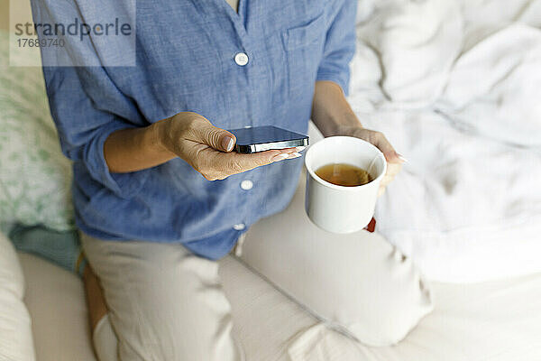 Hands of woman holding mobile phone and tea cup sitting on bed