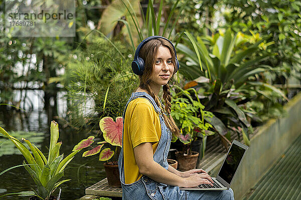 Junge Frau sitzt mit Laptop und hört Musik über kabellose Kopfhörer im Garten